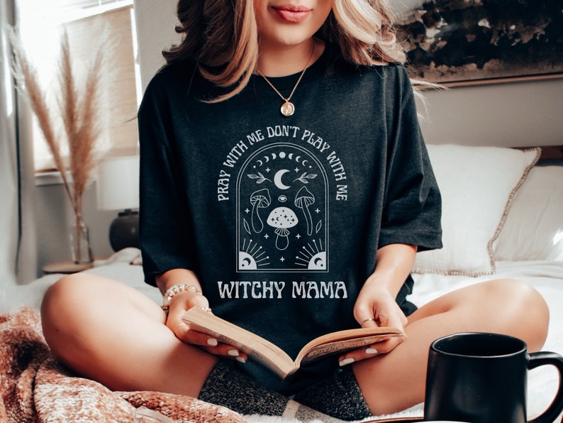 May include: A black t-shirt with a white graphic design featuring a crescent moon, stars, mushrooms, and the text "Pray with me don't play with me Witchy Mama".