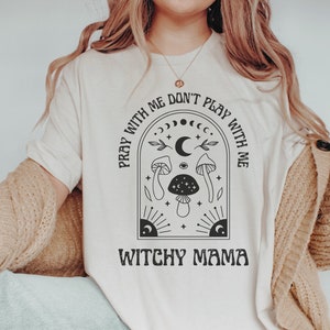 May include: A white t-shirt with a black graphic design featuring a crescent moon, stars, mushrooms, and the text "Pray with me don't play with me Witchy Mama".