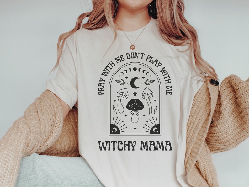 May include: A white t-shirt with a black graphic design featuring a crescent moon, stars, mushrooms, and the text "Pray with me don't play with me Witchy Mama".