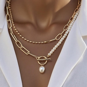 May include: A multi-strand gold-tone necklace with chain links, beads, and pearls. The necklace includes a toggle clasp and a dangling pearl. The jewellery is worn against a white collared shirt.