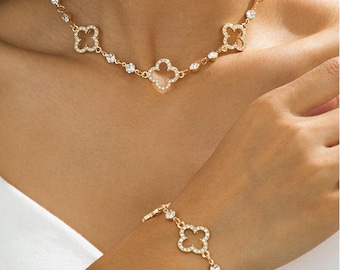 May include: Gold-toned jewelry set featuring a necklace and bracelet. The necklace and bracelet showcase a clover design with clear, sparkling accents. The jewelry is worn on a person, with the necklace around the neck and the bracelet on the wrist.