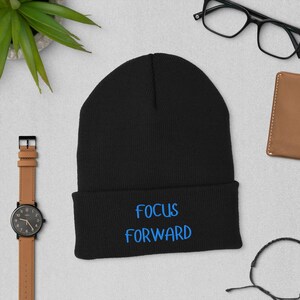 Pode incluir: Um gorro preto com as palavras "FOCUS FORWARD" bordadas em azul. O gorro é dobrado na aba. Outros itens na imagem incluem um relógio, óculos, uma carteira e uma planta.
