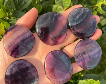 Puede incluir: Seis piedras de fluorita lisas, redondas y pulidas en tonos de morado, verde y rosa. Las piedras están dispuestas en una mano, con la piedra más grande en el centro.