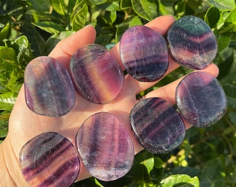 Puede incluir: Una colección de ocho piedras de fluorita pulidas, cada una con un patrón único de rayas púrpuras, verdes y amarillas. Las piedras son lisas y de forma ovalada, y están dispuestas en una mano.