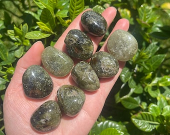 May include: A collection of tumbled green stones, varying in shades from olive to dark green, are held in an open hand. The stones have a smooth, polished appearance and are displayed against a backdrop of green foliage, with sunlight illuminating the scene.