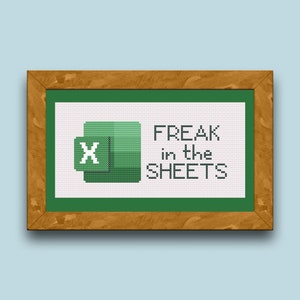 May include: Cross-stitch artwork in a wooden frame. The design features the Microsoft Excel logo in green and white, with the phrase "FREAK in the SHEETS" in black text. The background is white, and the frame is a warm brown.