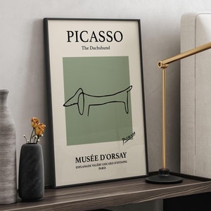 Könnte beinhalten: Gerahmter Kunstdruck mit einer Picasso-Zeichnung eines Dackels, Text "PICASSO The Dachshund" und "MUSÉE D'ORSAY". Eine goldfarbene Schreibtischlampe mit schwarzem Sockel steht auf einem Nachttisch aus dunklem Holz.