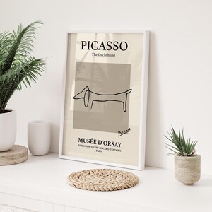 Pode incluir: Impressão de arte emoldurada com um desenho de Picasso de um dachshund. A impressão tem uma paleta de cores neutras com o texto "PICASSO" e "MUSÉE D'ORSAY". A obra de arte é exibida em uma superfície branca com plantas e um porta-copos.