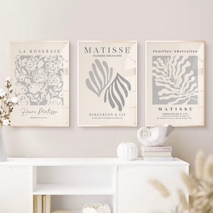 Puede incluir: Tres impresiones artísticas enmarcadas con fondos beige. Las impresiones presentan diseños florales y abstractos en gris, con texto como "LA ROSERAIE", "MATISSE" y "Feuilles Abstraites". Los marcos son de color madera clara.
