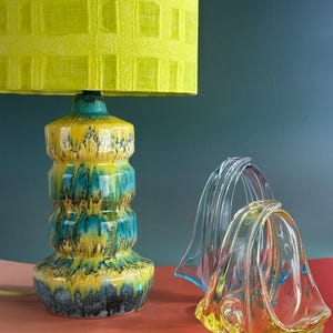 Könnte beinhalten: Eine lebendige Tischlampe mit gelbem, strukturiertem Lampenschirm steht auf einem Keramiksockel mit gelber, türkisfarbener und schwarzer Glasur. Zwei Glasskulpturen in klaren und bernsteinfarbenen Farbtönen stehen neben der Lampe.