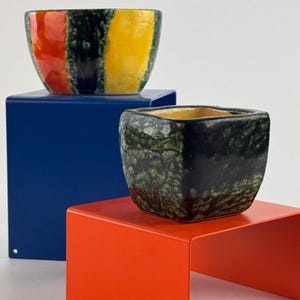 Può includere: Una ciotola in ceramica rotonda con strisce verticali rosse, nere e gialle posata su un supporto in metallo blu. Accanto, un vaso quadrato in ceramica con una smaltatura maculata verde scuro e nera poggia su un supporto in metallo arancione.
