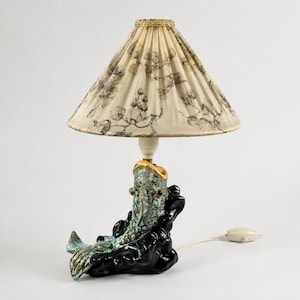 Könnte beinhalten: Eine Tischlampe mit einem Keramikfuß in Form eines blaugrünen und schwarzen Fisches auf einem dunklen Sockel. Der konische, plissierte cremeweiße Lampenschirm ist mit einem schwarzen botanischen Muster und einem goldenen Rand verziert.