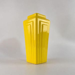 Puede incluir: Un florero de cerámica de color amarillo brillante con acabado lustroso, que presenta una forma geométrica angular con paneles verticales empotrados que se estrechan hacia la base y una abertura superior cuadrada y plana.