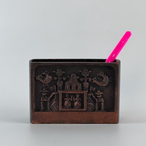 May include: A rectangular bronze-toned metal desk organizer featuring a relief design of two figures standing in a central structure flanked by stylized birds and floral motifs. A single bright neon pink pen is inserted into the open top compartment.