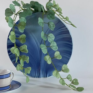Puede incluir: Un jarrón de pared azul decorativo con un patrón arremolinado, que sostiene vides verdes con hojas. Una pequeña taza de té y un platillo con rayas azules son visibles en primer plano, sugiriendo un tema de decoración del hogar.