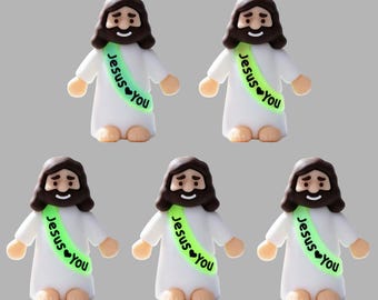 May include: Five identical small figurines of a person with brown hair and a beard, wearing a white robe and a light green sash. The sash features the black text "Jesus ♥ you" printed on it. Each figurine has a simple, stylized design.