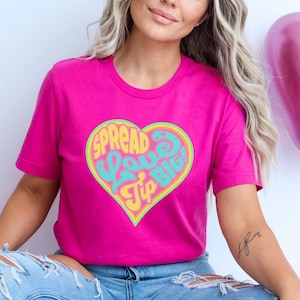 May include: A magenta t-shirt featuring a heart-shaped graphic with the words "Spread Love Big Tip!" in a retro font. The heart has a yellow and light blue outline. The person is wearing light blue ripped jeans and white sneakers.