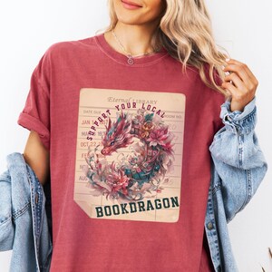 May include: A crimson short-sleeve t-shirt featuring a graphic of a dragon intertwined with flowers and books on a library checkout card background. The text reads "SUPPORT YOUR LOCAL BOOKDRAGON" and "Eternal LIBRARY" in dark lettering.