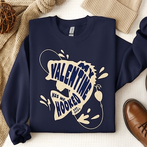 May include: Navy blue sweatshirt with a graphic of a fish with the text "My Valentine Has Me Hooked Est. 2024" in a cream color.