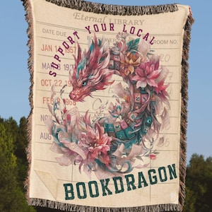 May include: A cream-colored throw blanket with a dragon design. The dragon is pink, red, and teal, surrounded by flowers and books. Text includes "Eternal Library," "Support Your Local," and "Bookdragon."