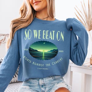Comfort Colors 6014 long sleeve tee in Blue Jean — So We Beat On Fitzgerald quote long sleeve with retro-illustrated moonlit harbor scene and 1925–2025 centennial detail. Lifestyle shot, model with arm raised. Great gift for Fitzgerald fans, book nerds, and literary apparel lovers celebrating 100 years of The Great Gatsby.