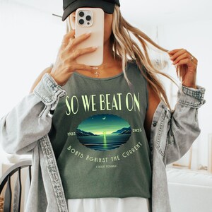 May include: A dark olive green tank top featuring the text "SO WE BEAT ON" in light yellow, with a central oval graphic of a sunset over water, the years "1925" and "2025", the text "BOATS AGAINST THE CURRENT", and "F. SCOTT FITZGERALD".
