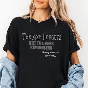 May include: Black t-shirt with the text "The Axe Forgets But The Neck Remembers" in a serif font. Below the quote is "Thomas Cromwell (Probably)". The shirt has rolled-up sleeves.