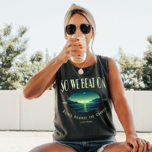May include: A charcoal grey sleeveless tank top featuring the text SO WE BEAT ON and BOATS AGAINST THE CURRENT in light green lettering. A central oval graphic depicts a sunset over water with the years 1925 and 2025 and F Scott Fitzgerald.