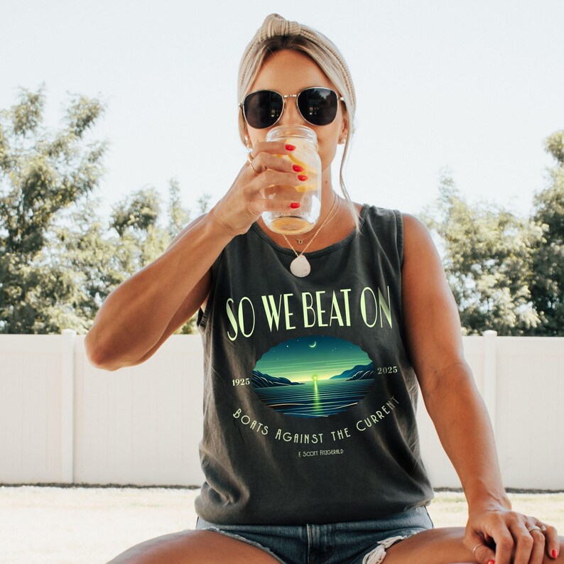 May include: A charcoal grey sleeveless tank top featuring the text SO WE BEAT ON and BOATS AGAINST THE CURRENT in light green lettering. A central oval graphic depicts a sunset over water with the years 1925 and 2025 and F Scott Fitzgerald.