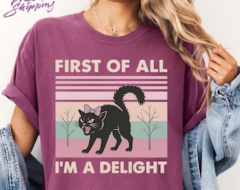 May include: A maroon Comfort Colors t-shirt with the text "FIRST OF ALL I'M A DELIGHT" above a graphic of a black cat with a pink bow. The cat is snarling with its mouth open. The shirt also has the words "Free Shipping" in cursive.