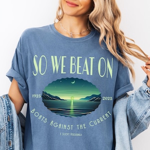 Comfort Colors 1717 classic tee in Blue Jean — So We Beat On Great Gatsby quote design with retro moonlit bay illustration and Boats Against the Current arc. Lifestyle shot, model wearing denim jacket. Perfect literary gift for book lovers, Fitzgerald fans, and BookTok aesthetic seekers who love classic lit graphic tees.