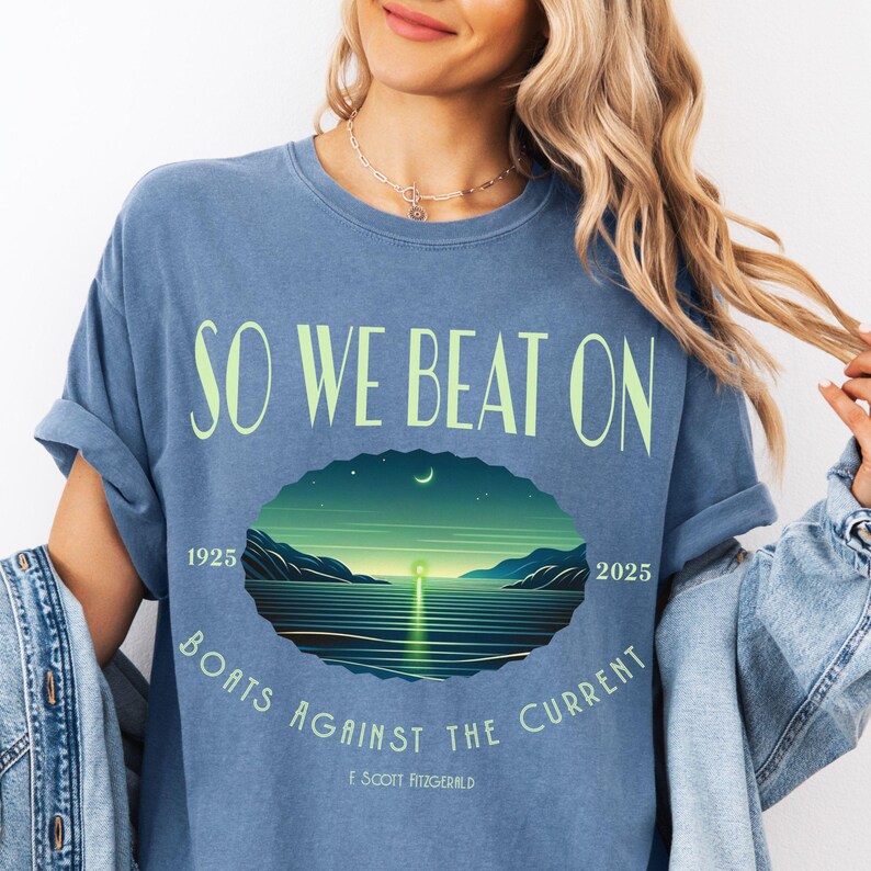Comfort Colors 1717 classic tee in Blue Jean — So We Beat On Great Gatsby quote design with retro moonlit bay illustration and Boats Against the Current arc. Lifestyle shot, model wearing denim jacket. Perfect literary gift for book lovers, Fitzgerald fans, and BookTok aesthetic seekers who love classic lit graphic tees.