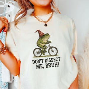 May include: Cream-colored t-shirt featuring a whimsical illustration of a green frog riding a bicycle and wearing a brown and red polka-dot hat. The text "DON'T DISSECT ME, BRUH!" is printed below the frog. The shirt has a relaxed fit.