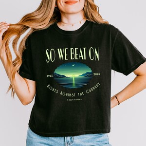 May include: A black short-sleeve t-shirt featuring the text SO WE BEAT ON and BOATS AGAINST THE CURRENT in light green. A central oval graphic depicts a sunset over water with the years 1925 and 2025 and the name F. Scott Fitzgerald printed below.