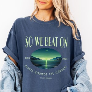 Comfort Colors 1717 classic tee in Denim — So We Beat On Great Gatsby quote graphic tee with crescent moon bay illustration. Lifestyle shot, model with sun pendant necklace. Great gift for Fitzgerald fans, book nerds, and literary apparel lovers celebrating 100 years of The Great Gatsby 1925–2025.