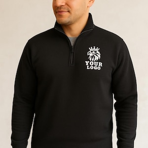 May include: Black quarter-zip sweatshirt with a white logo of an eagle wearing a crown and the text "YOUR LOGO". The sweatshirt has a high collar and a silver zipper. The model is wearing a gray undershirt.