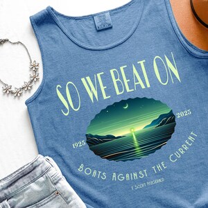 May include: A blue sleeveless tank top featuring the text SO WE BEAT ON and BOATS AGAINST THE CURRENT with the name F. SCOTT FITZGERALD. A central graphic depicts a night landscape with a glowing light over water, the years 1925 and 2025.