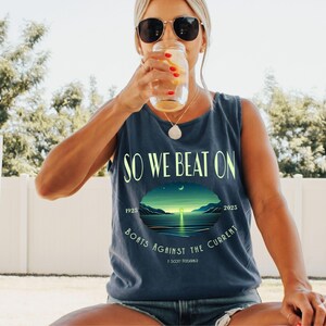 May include: A dark blue sleeveless tank top featuring the text SO WE BEAT ON and BOATS AGAINST THE CURRENT in light green lettering. A central graphic depicts a sunset over a calm body of water with the years 1925 and 2025 and F. Scott Fitzgerald.