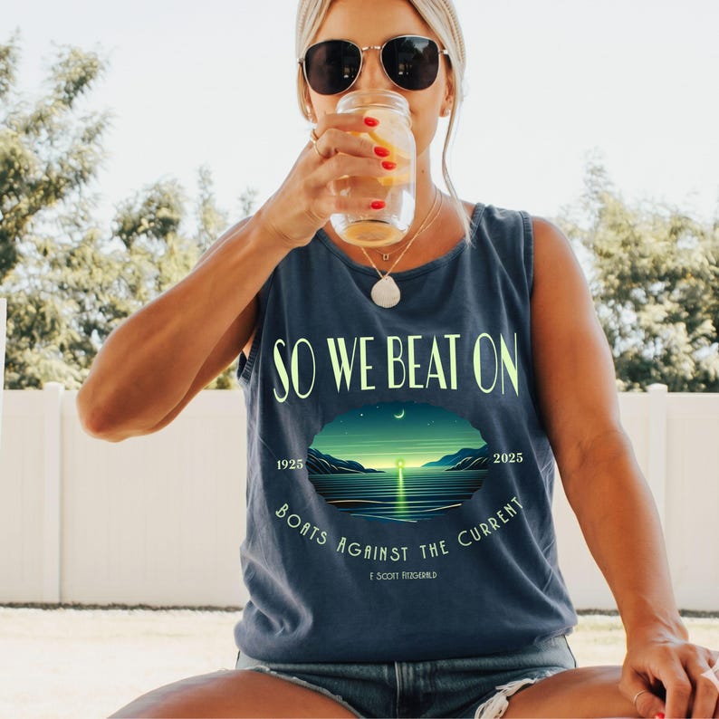May include: A dark blue sleeveless tank top featuring the text SO WE BEAT ON and BOATS AGAINST THE CURRENT in light green lettering. A central graphic depicts a sunset over a calm body of water with the years 1925 and 2025 and F. Scott Fitzgerald.