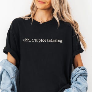 May include: Black t-shirt with the phrase "Shh... I'm plot twisting" in white lettering. The shirt has short sleeves and a relaxed fit. The model is wearing a denim jacket.