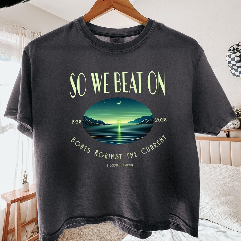 May include: A charcoal grey t-shirt featuring the text SO WE BEAT ON and BOATS AGAINST THE CURRENT in light yellow. A central oval graphic depicts a sunset over water with the years 1925 and 2025, and the name F. Scott Fitzgerald below.