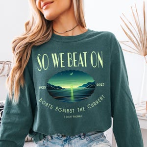 Comfort Colors 6014 long sleeve tee in Blue Spruce — So We Beat On Great Gatsby quote design with retro moonlit bay illustration and Boats Against the Current arc. Lifestyle shot, model with black choker necklace. Literary gift for book lovers, Fitzgerald fans, and BookTok aesthetic seekers who love classic lit long sleeve tees.