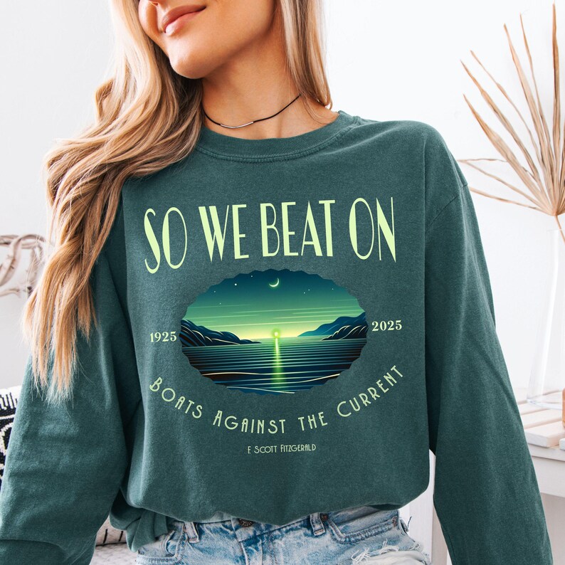 Comfort Colors 6014 long sleeve tee in Blue Spruce — So We Beat On Great Gatsby quote design with retro moonlit bay illustration and Boats Against the Current arc. Lifestyle shot, model with black choker necklace. Literary gift for book lovers, Fitzgerald fans, and BookTok aesthetic seekers who love classic lit long sleeve tees.