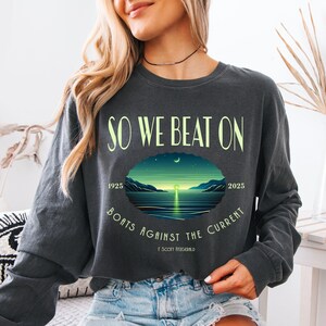 Comfort Colors 6014 long sleeve tee in Pepper — So We Beat On Fitzgerald quote long sleeve with retro moonlit bay illustration and Boats Against the Current arc. Lifestyle shot, model in denim shorts. For bookish aesthetic lovers, Fitzgerald fans, and readers celebrating 100 years of The Great Gatsby 1925–2025.