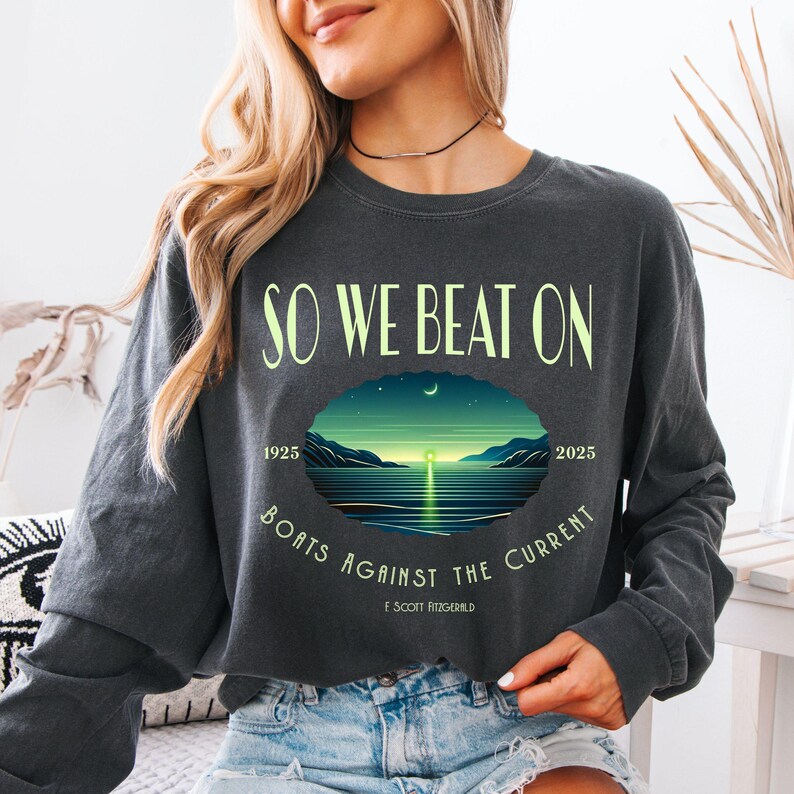 Comfort Colors 6014 long sleeve tee in Pepper — So We Beat On Fitzgerald quote long sleeve with retro moonlit bay illustration and Boats Against the Current arc. Lifestyle shot, model in denim shorts. For bookish aesthetic lovers, Fitzgerald fans, and readers celebrating 100 years of The Great Gatsby 1925–2025.