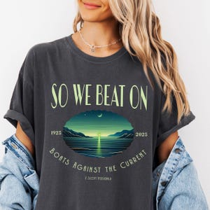 Comfort Colors 1717 classic tee in Pepper — So We Beat On Great Gatsby quote tee with neon green moonlit bay and Boats Against the Current arc. Lifestyle shot, model layered with cream cardigan and gold chain necklace. For bookish aesthetic lovers, Fitzgerald fans, and readers celebrating 100 years of The Great Gatsby.