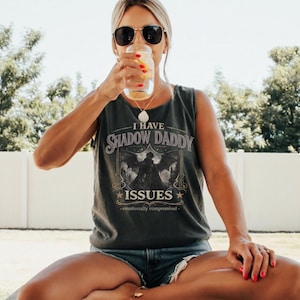 May include: Dark gray tank top with the text "I Have Shadow Daddy Issues" and a graphic of a bat-winged figure. The tank top is paired with denim shorts. The person is wearing sunglasses and drinking from a glass.