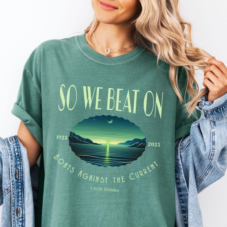 Comfort Colors 1717 classic tee in Light Green — So We Beat On Fitzgerald quote tee with retro-illustrated glowing harbor scene. Lifestyle shot, model in denim jacket. For BookTok style, reader girl summer aesthetic, and classic lit lovers who wear their favorite quotes and want a unique Great Gatsby centennial gift.