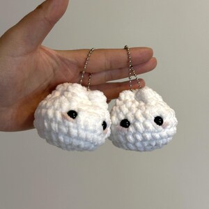 Puede incluir: Dos llaveros blancos con forma de nube, tejidos a ganchillo, con ojos negros y mejillas rosas. Cada llavero está sujeto a una cadena plateada. Los llaveros se sostienen sobre un fondo liso.