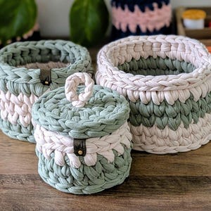Può includere: Un set di tre cesti portaoggetti rotondi fatti a mano, nei toni del verde e bianco sporco. I cesti sono realizzati in tessuto spesso intrecciato e presentano un accento in pelle. Un cesto ha un coperchio con manico intrecciato.